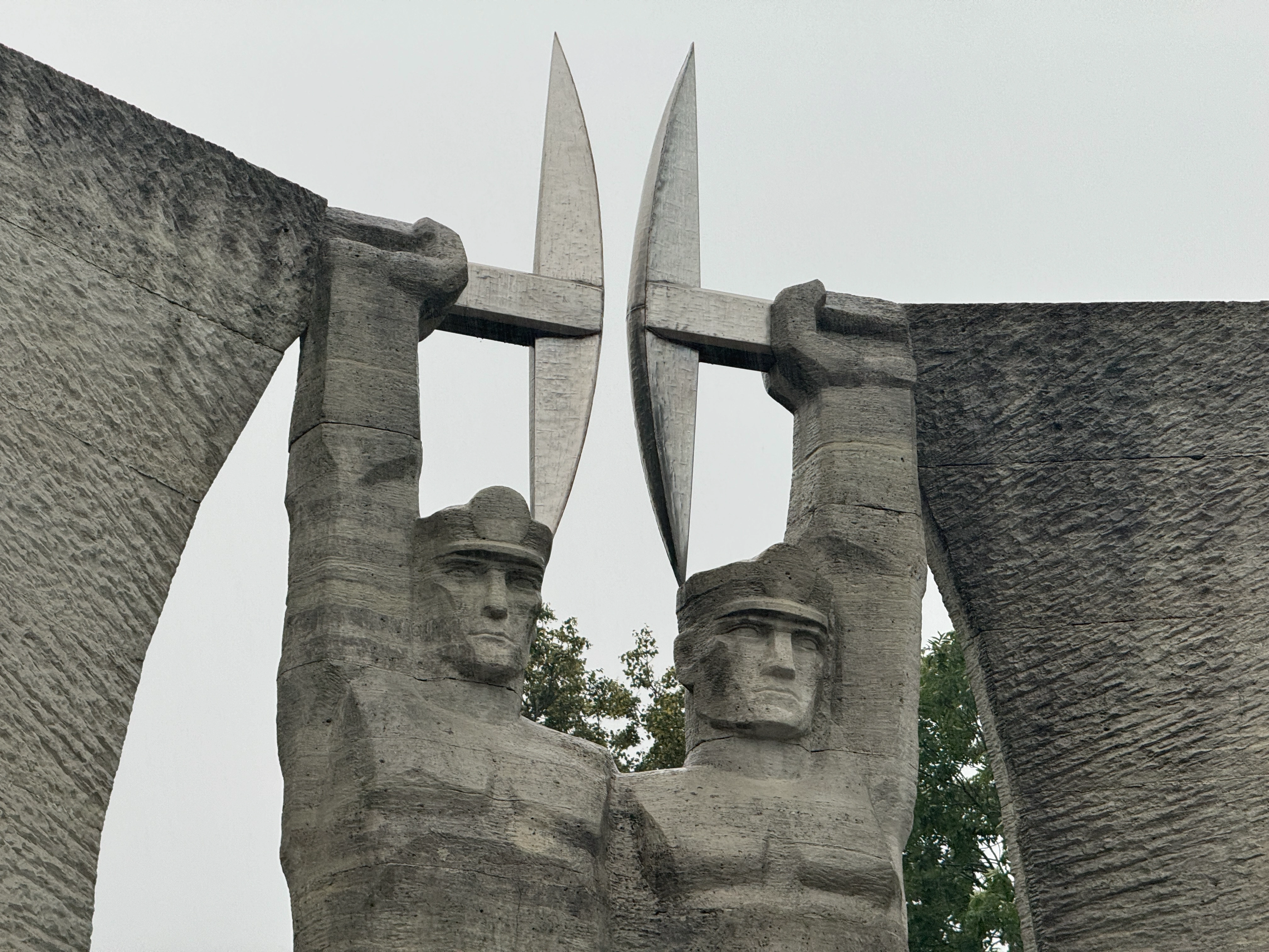 Angled close-up from the left of the brutalist concrete Glory to Work monument in Kohtla-Järve, detailing the profiles, upper bodies, and raised pickaxes of the two sculpted miners.