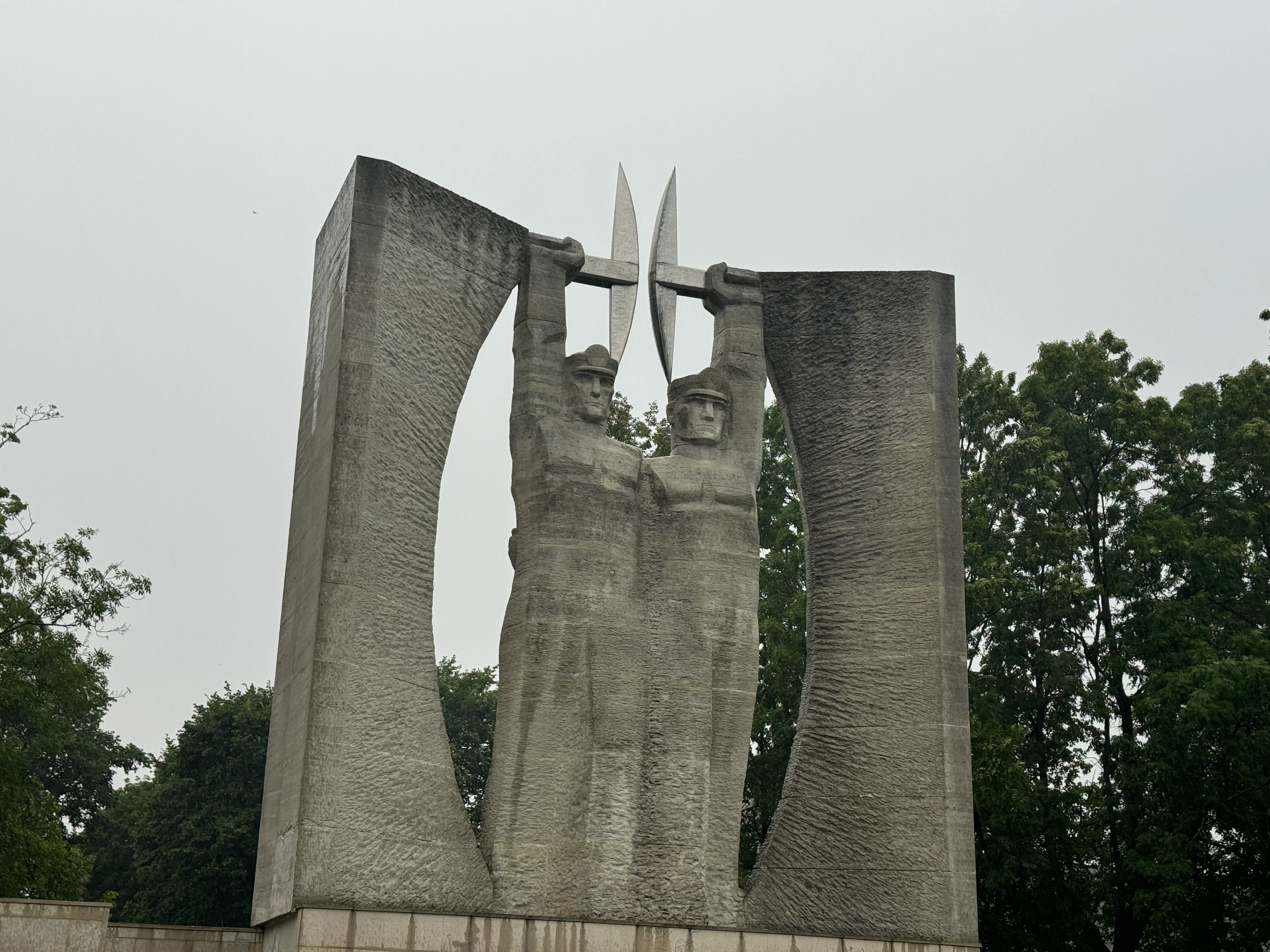 Wide view of the brutalist concrete Glory to Work monument in Kohtla-Järve. Two sculpted miners hold pickaxes aloft between massive curved pillars on a stepped stone base.