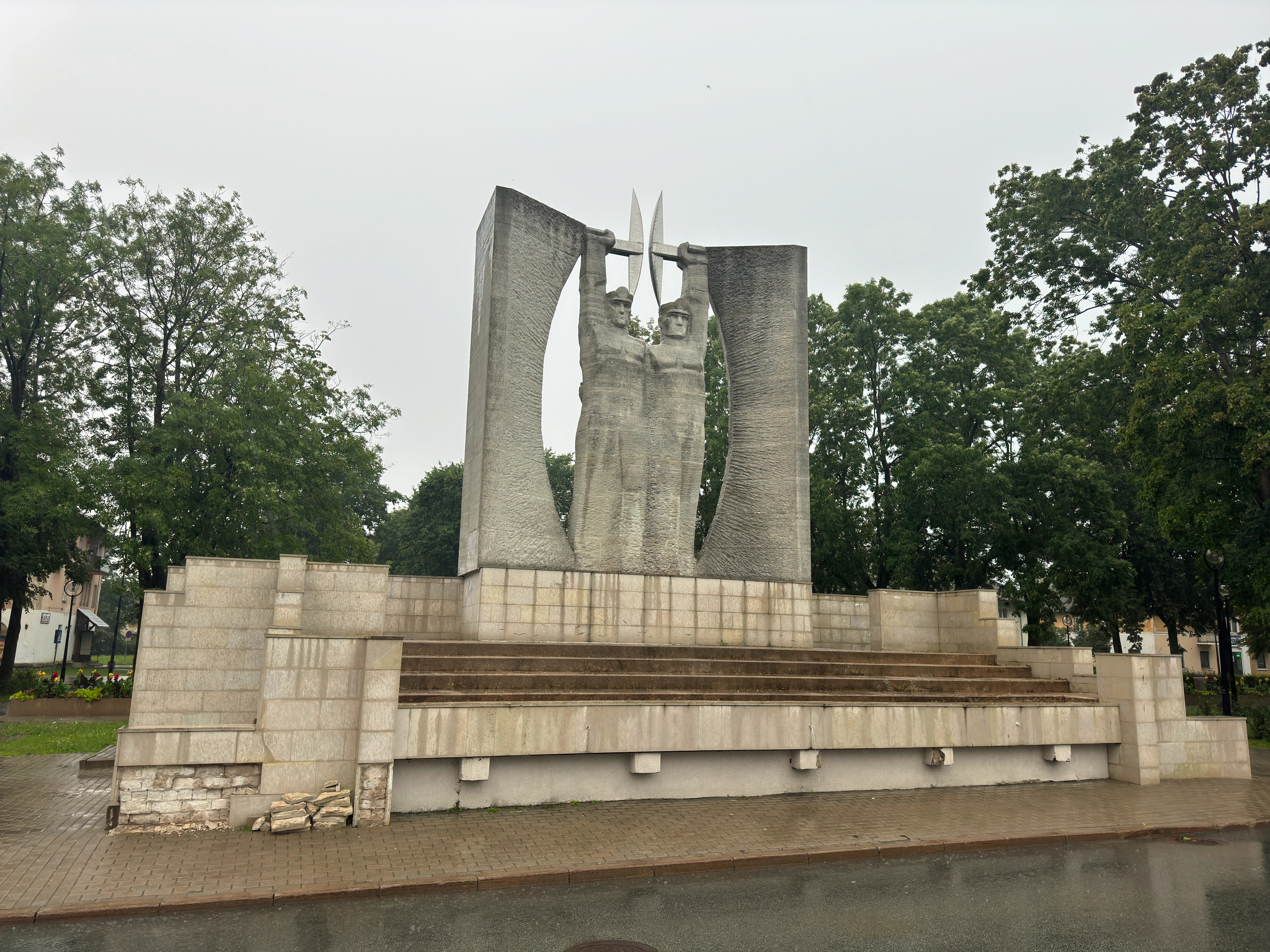 Wide angled view from the left of the brutalist concrete Glory to Work monument in Kohtla-Järve, featuring two miners holding pickaxes between large curved pillars on a stepped stone base.