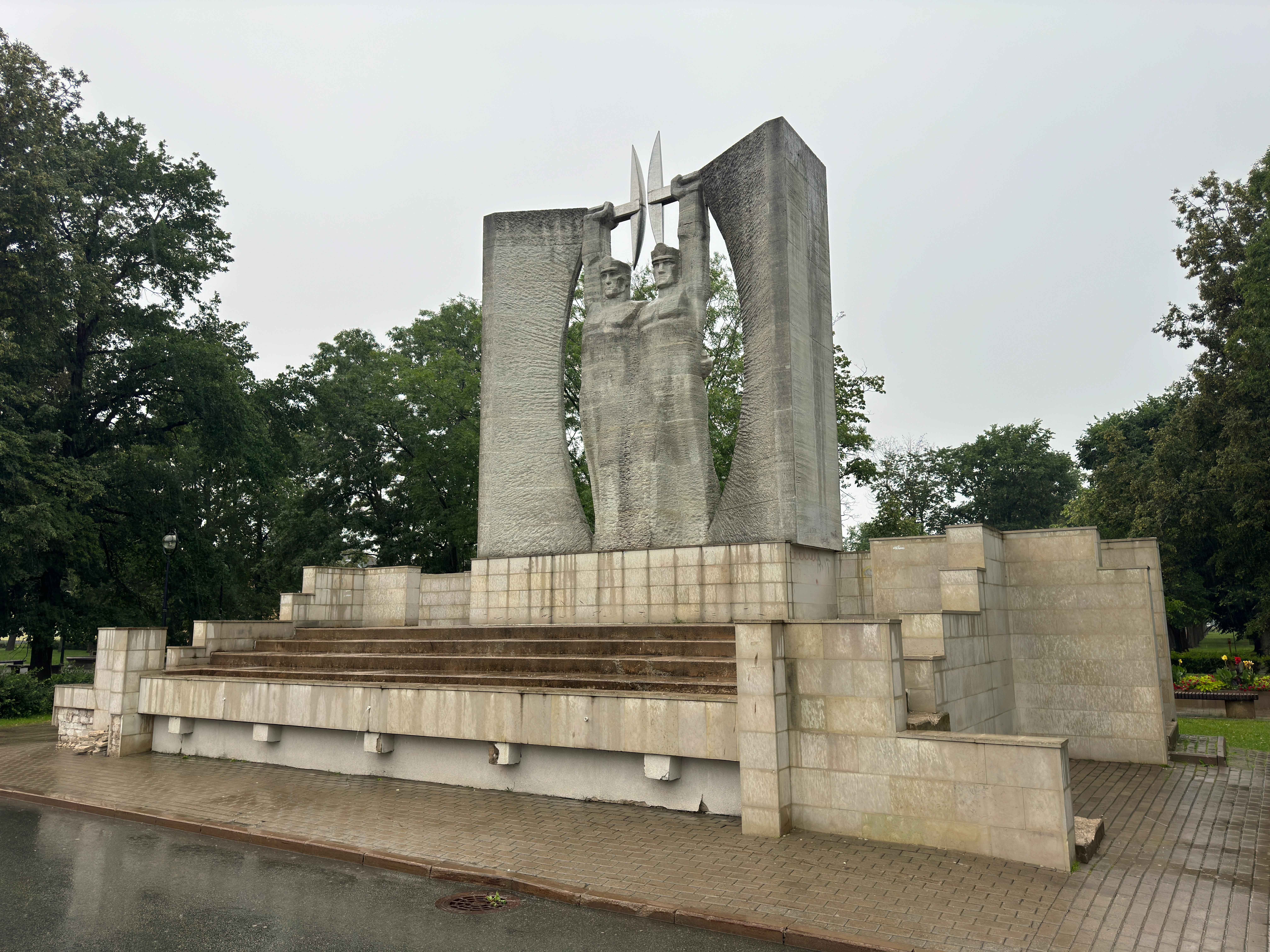 Wide angled view from the right of the brutalist concrete Glory to Work monument in Kohtla-Järve, featuring two miners holding pickaxes between large curved pillars on a stepped stone base.