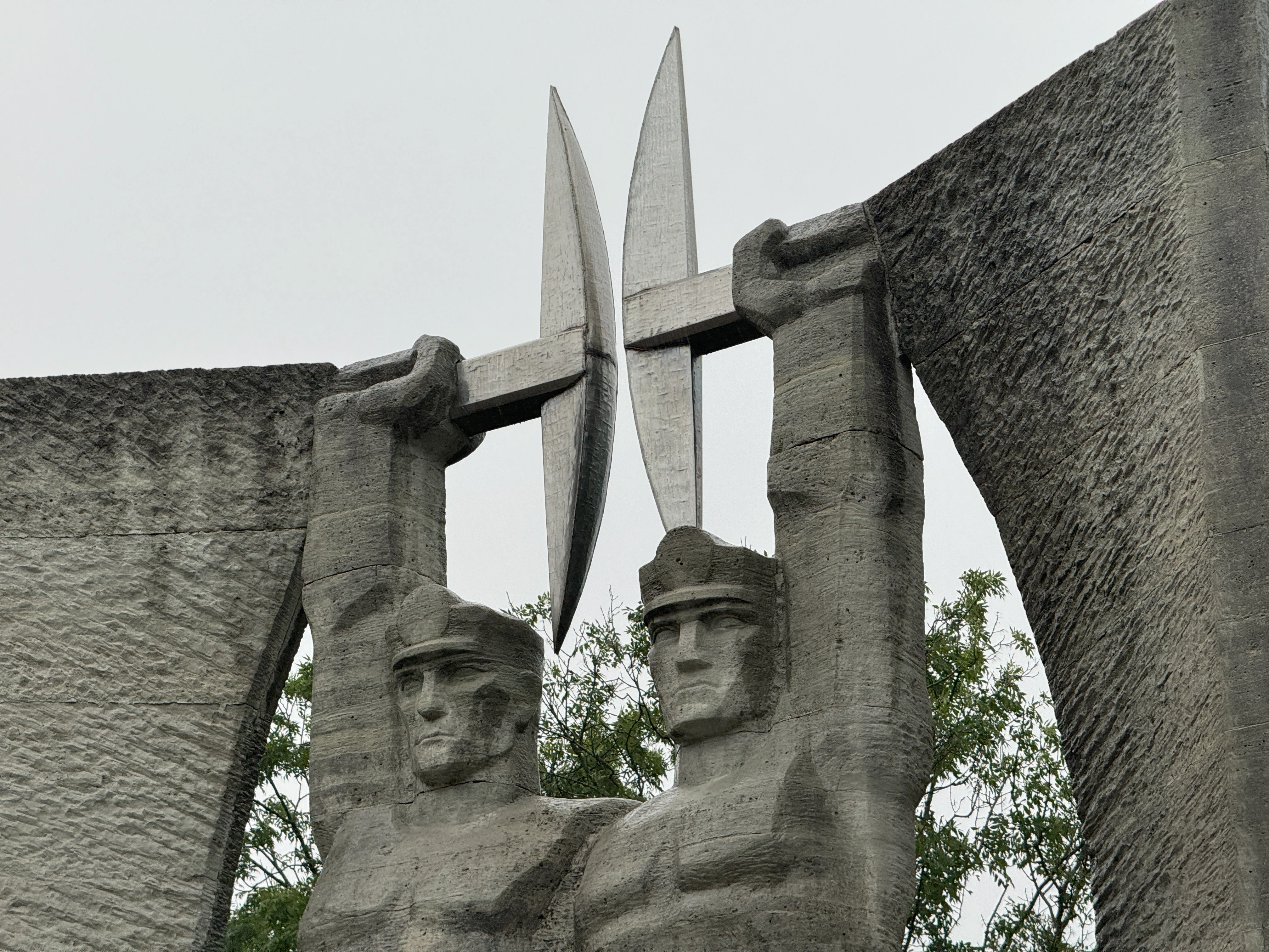 Angled close-up from the right of the brutalist concrete Glory to Work monument in Kohtla-Järve, detailing the profiles, upper bodies, and raised pickaxes of the two sculpted miners.