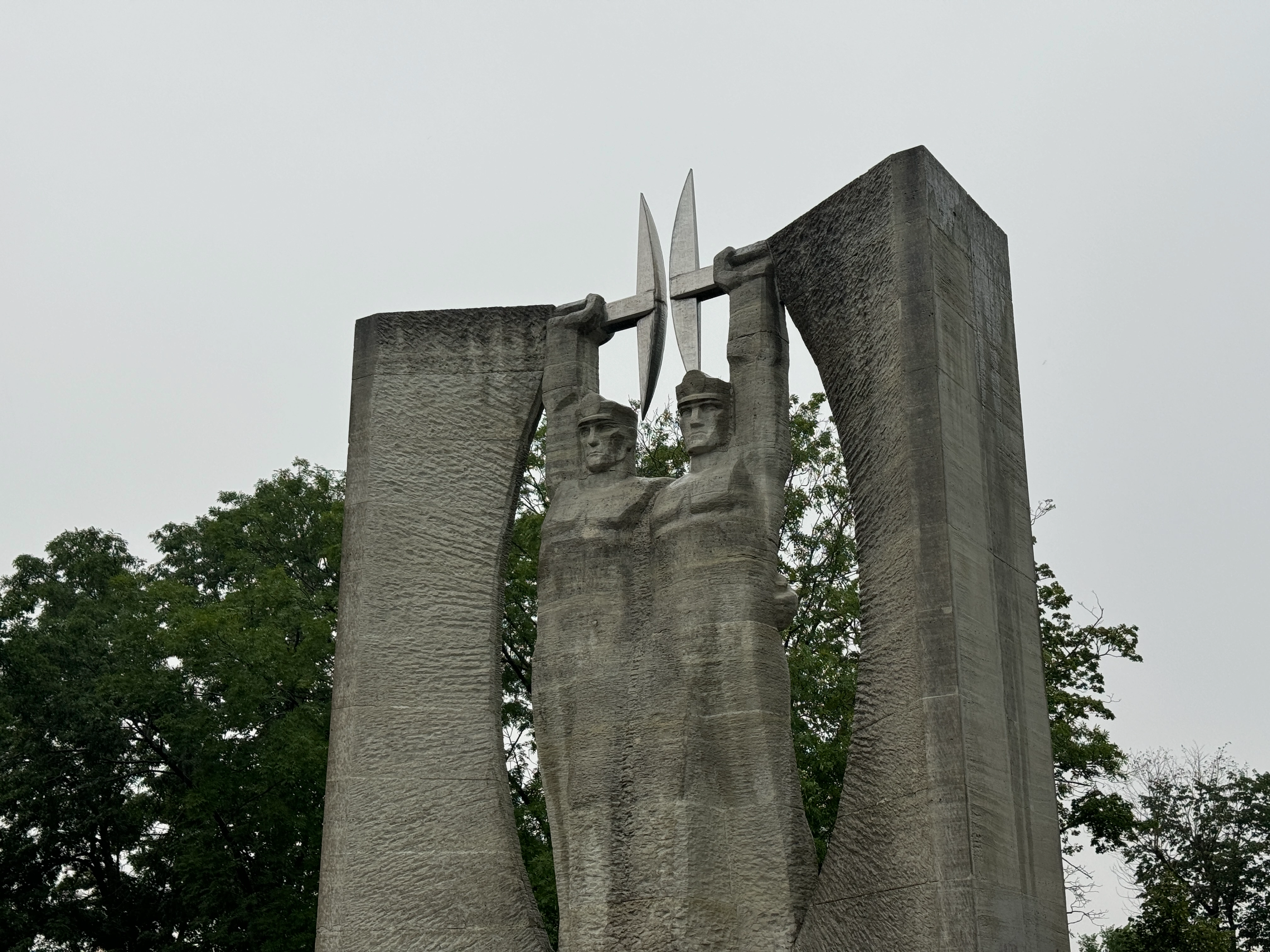 Low angle side view of the brutalist concrete Glory to Work monument in Kohtla-Järve, showing the side profile of the sculpted miners and the thick curved concrete pillar.