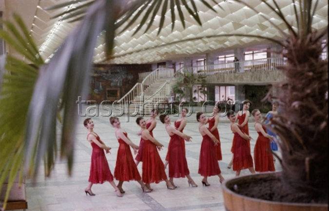Black and white historical photography of the spacious modernist interior main foyer of the Republican Youth Center named after Yuri Gagarin in Chisinau during the 1970s, showcasing a futuristic three-dimensional geometric ceiling, an elegant open staircase, the prominent large dark mural artwork on the back wall, and groups of people mingling and sitting at tables.