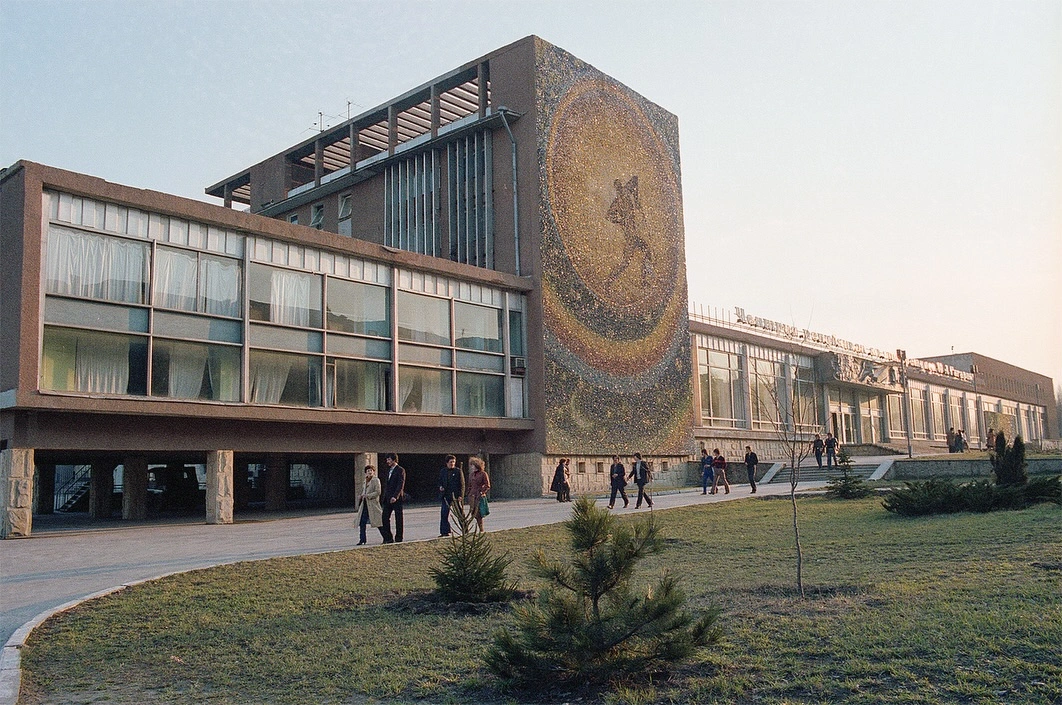 Wide exterior architectural photograph of the Republican Youth Center named after Yuri Gagarin in Chisinau, Moldova from April 1982. The image highlights the Soviet modernist design, large panoramic glass facade windows, concrete support pillars, and the massive "Plowman of the Universe" space-themed mosaic by artist Aurel David on the prominent side wall, with pedestrians walking along the path in the foreground.