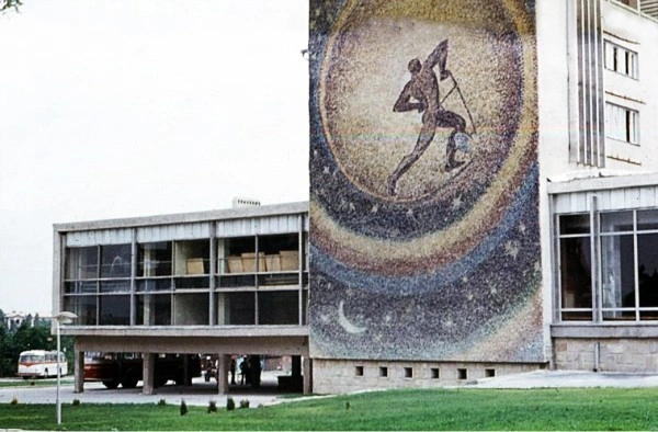 Close-up exterior color photograph from 1972 highlighting the towering "Plowman of the Universe" mosaic by Aurel David on the side wall of the Gagarin Youth Center in Chisinau, showing a running figure against a starry space background, with a red Sputnik tourist bus parked in the shade underneath the building's concrete overhang.