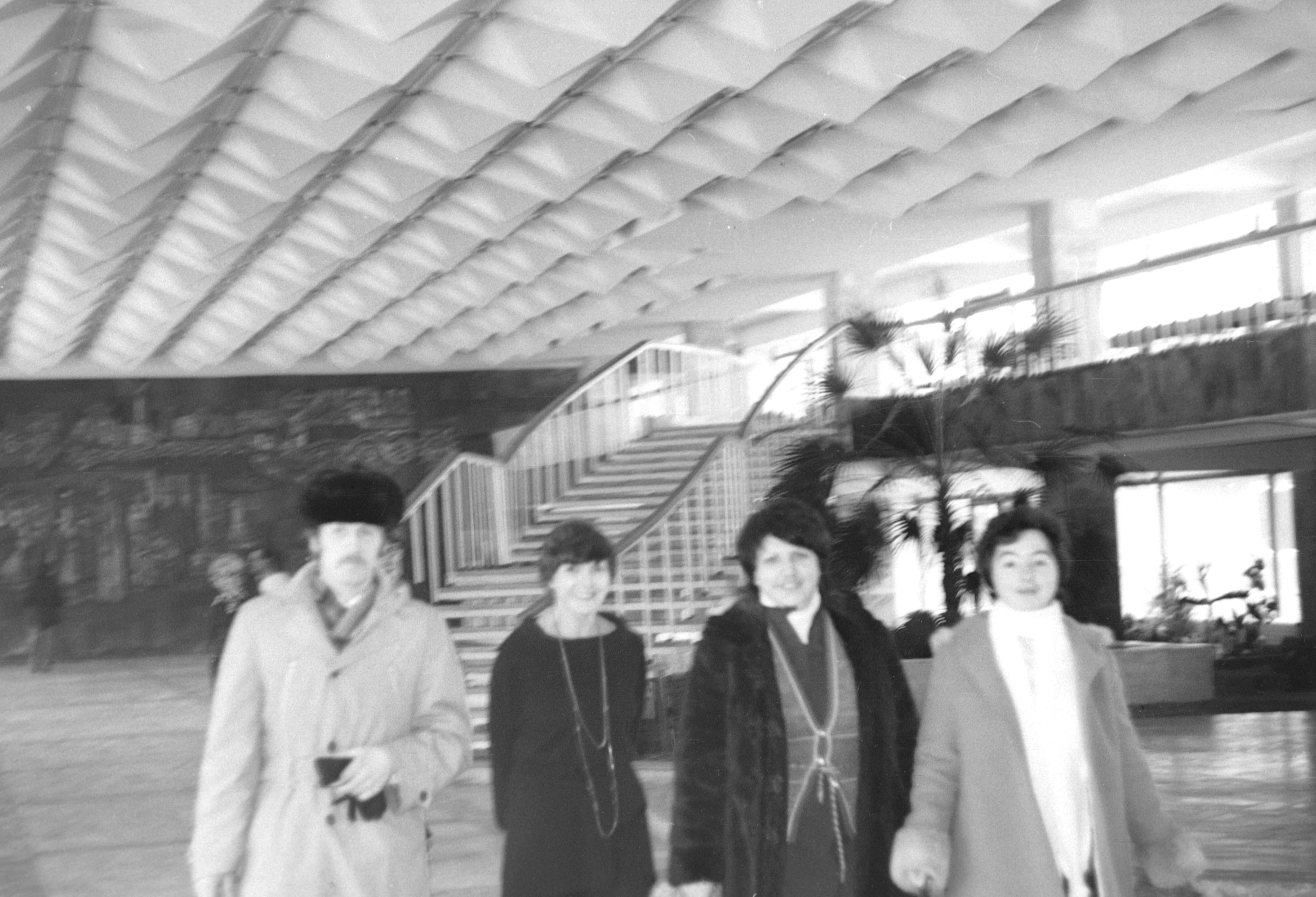 Vintage 1983 black and white photograph capturing famous Soviet poet Bella Akhmadulina and three other individuals posing inside the brightly lit main foyer of the Gagarin Youth Center in Chisinau, standing in front of the large dark mural artwork on the back wall, with the distinctive open staircase serving as the backdrop.