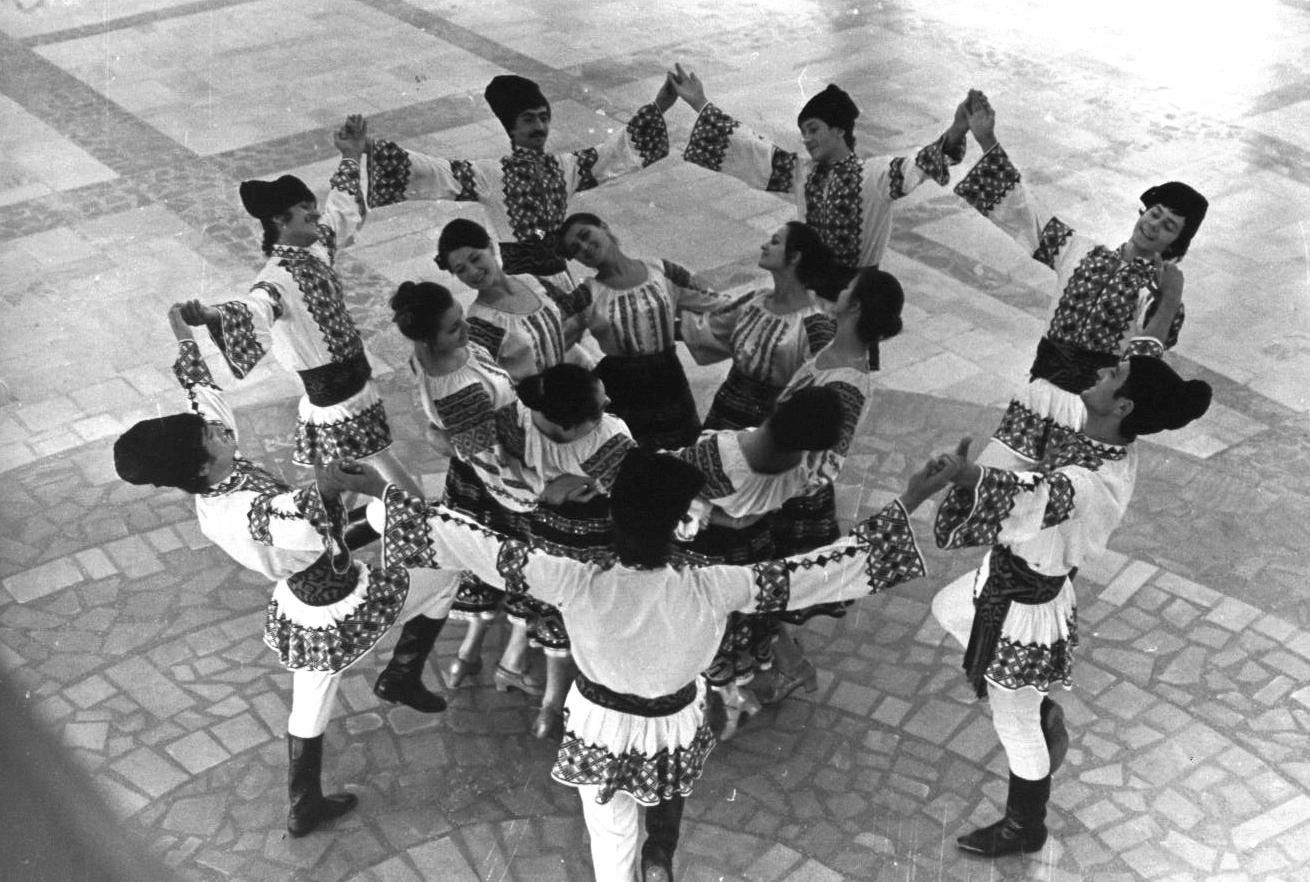 Dynamic high-angle black and white photograph from 1974 showing an amateur Moldovan folk dance ensemble holding hands in a circular star formation on the tiled marble floor of the Gagarin Youth Center, featuring male and female dancers wearing elaborately embroidered traditional national costumes and tall black hats.