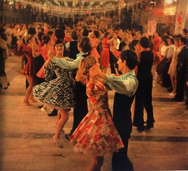 Warmly lit vintage 1980 color photograph of a massive festive ballroom dance event taking place inside the main hall of the Gagarin Youth Center in Chisinau, filled with dozens of young couples in retro clothing dancing together on the marble floor under festive party garlands strung across the room's unique modern ceiling.