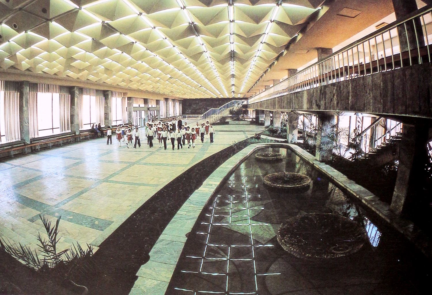 Expansive wide-angle color photograph from 1984 capturing the massive scale of the Gagarin Youth Center's main foyer viewed from the stage side. It showcases the long indoor reflecting pools in the foreground, a group of children walking across the wide tiled floor, and a large dark artwork mural on the far back wall under the brightly illuminated three-dimensional geometric ceiling.