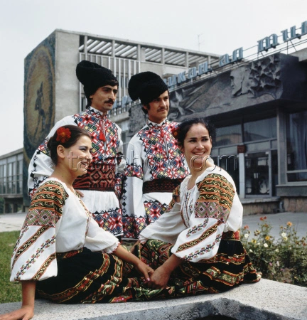 Color group portrait from September 1974 featuring four members of the Moldavian Republican Youth Center folk ensemble wearing intricately embroidered traditional costumes and tall black hats, sitting outside the main entrance of the building beneath the large dark bas-relief sculpture mounted above the doors.