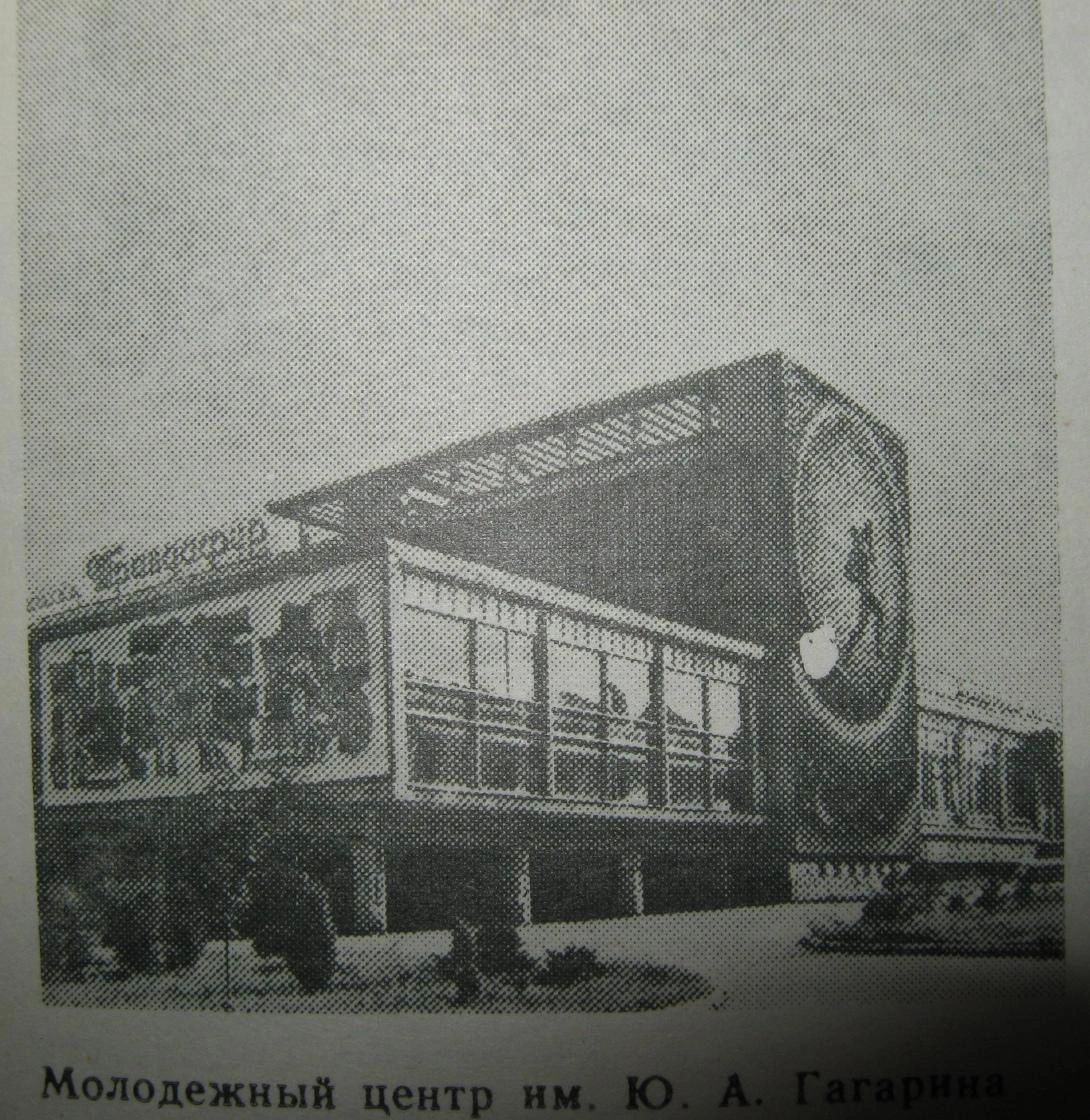 An early black and white historical press photograph of the lower left wing of the Gagarin Youth Center in Chisinau (the wing protruding above the parking lot). The photo shows the glass facade covered with a large dark, graphic panel or a temporary abstract mosaic/banner featuring abstract figures, complementing the main mosaic.