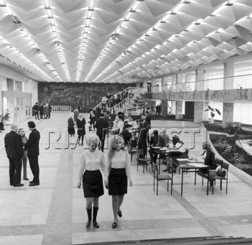 Black and white historical photography of the spacious modernist interior main foyer of the Republican Youth Center named after Yuri Gagarin in Chisinau during the 1970s, showcasing a futuristic three-dimensional geometric ceiling, an elegant open staircase, the prominent large dark mural artwork on the back wall, and groups of people mingling and sitting at tables.