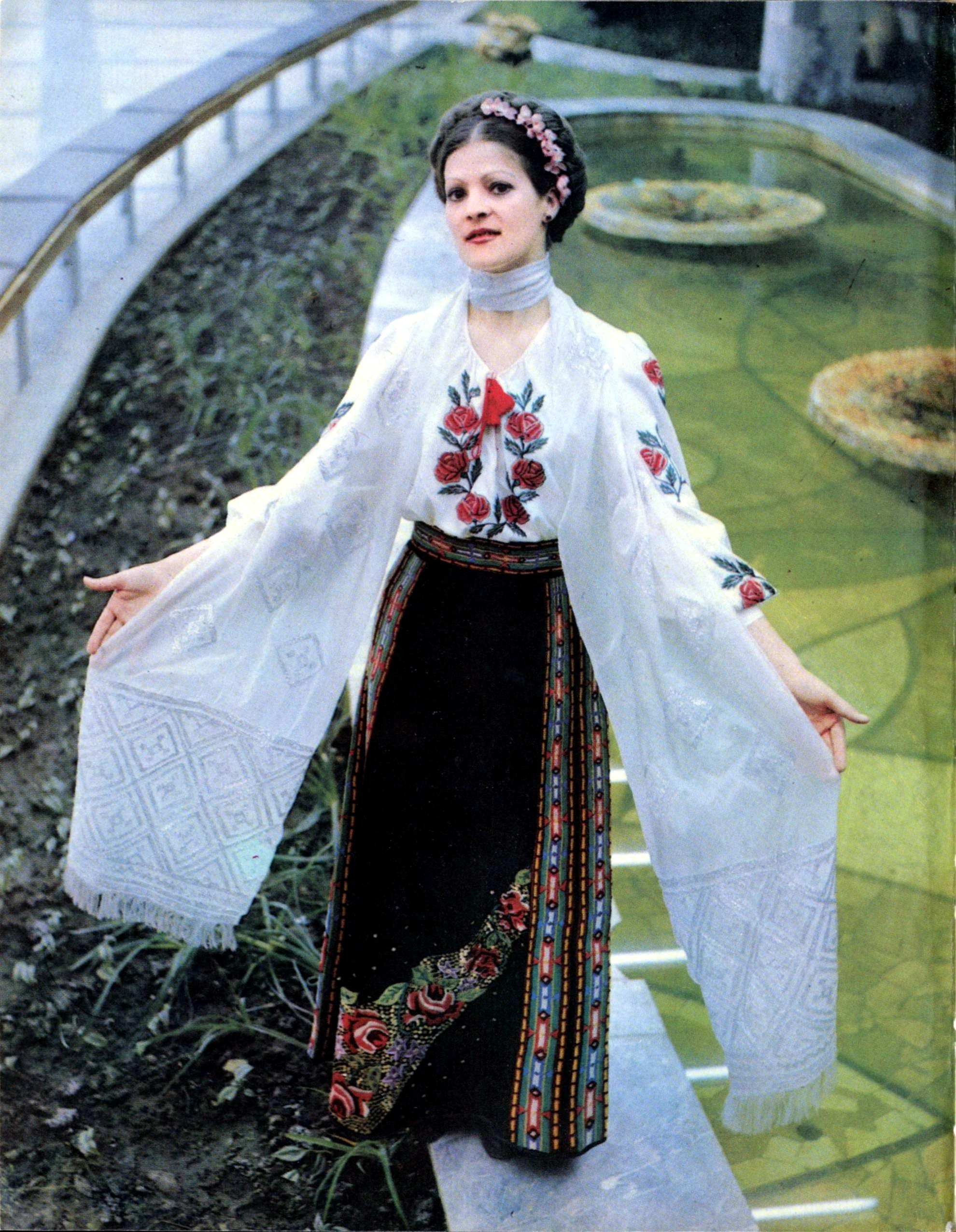 High-quality 1985 color photograph of a woman modeling a beautiful traditional Moldovan national costume featuring red rose embroidery, standing directly beside the indoor decorative water pools and fountains located within the main hall of the Gagarin Youth Center in Chisinau.