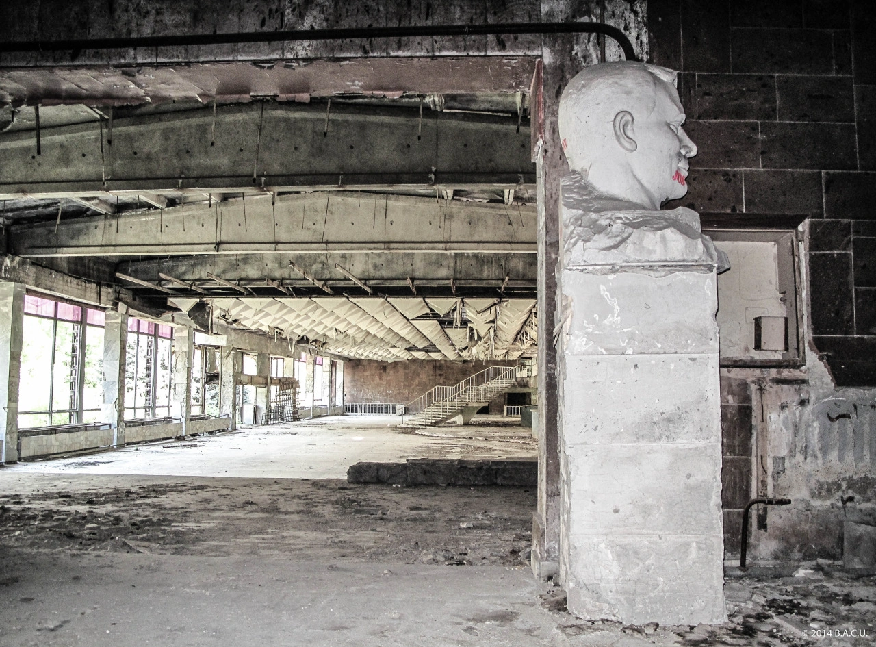 Post-apocalyptic style color photograph of the abandoned, ruined interior of the former Gagarin Youth Center in Chisinau, featuring a severely damaged and vandalized white stone bust of Yuri Gagarin with red graffiti resting on a concrete pillar in the foreground, with the collapsed geometric ceiling and debris-filled hall in the background.