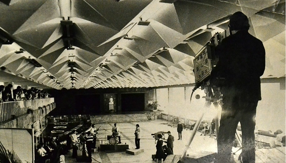 Historical black and white photograph showing a live television broadcast inside the grand hall of the Gagarin Youth Center. A cameraman with a large TV studio camera in the foreground is filming a live band performing in the middle of the room, while a raised podium featuring a prominent white bust of Yuri Gagarin stands in the center of the background.