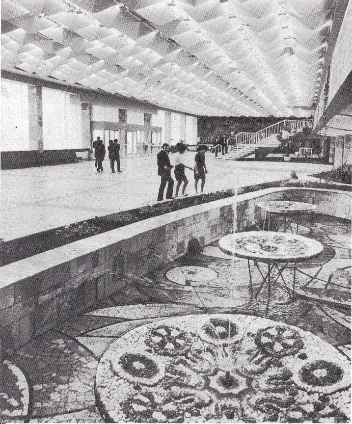 Black and white 1976 architectural interior photograph showcasing the decorative indoor water features of the Gagarin Youth Center in Chisinau, highlighting the mosaic-tiled circular fountain basins in the foreground, and looking across the hall toward the grand staircase and the large dark mural artwork on the back wall under the futuristic geometric ceiling.