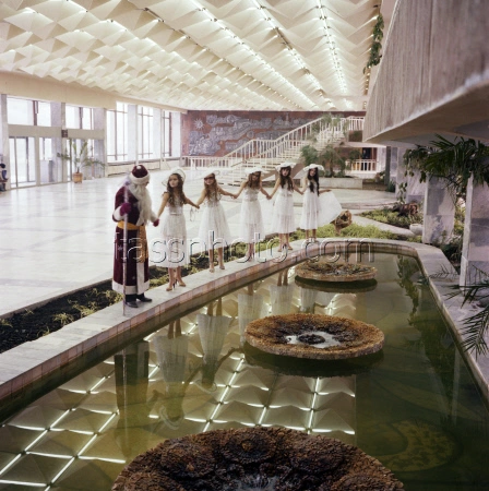 Historical black and white photograph, potentially from the 1980s or late 1970s, capturing a Christmas or New Year's celebration in the large foyer of the Republican Youth Center named after Yuri Gagarin in Chisinau. A group of young children in festive clothing, along with a man dressed as Ded Moroz (Soviet Santa Claus), are standing on the mosaic-inlaid circular edge of the indoor pools and fountains. In the background, the open staircase and part of the large dark mural artwork are visible.