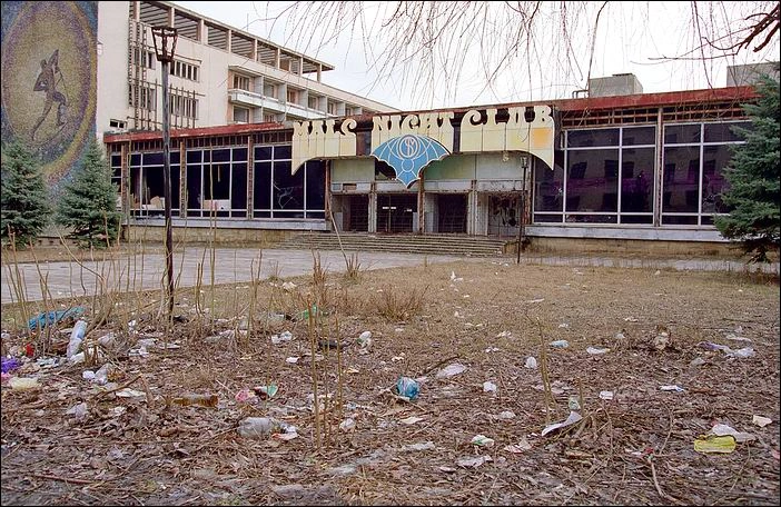 Color photograph from 2005 showing the dilapidated exterior of the closed MALS Night Club, formerly the Gagarin Youth Center in Chisinau, featuring a broken and weathered entrance sign with a blue bat logo, smashed glass windows, and a neglected front yard covered in overgrown weeds, dead branches, and scattered plastic litter.