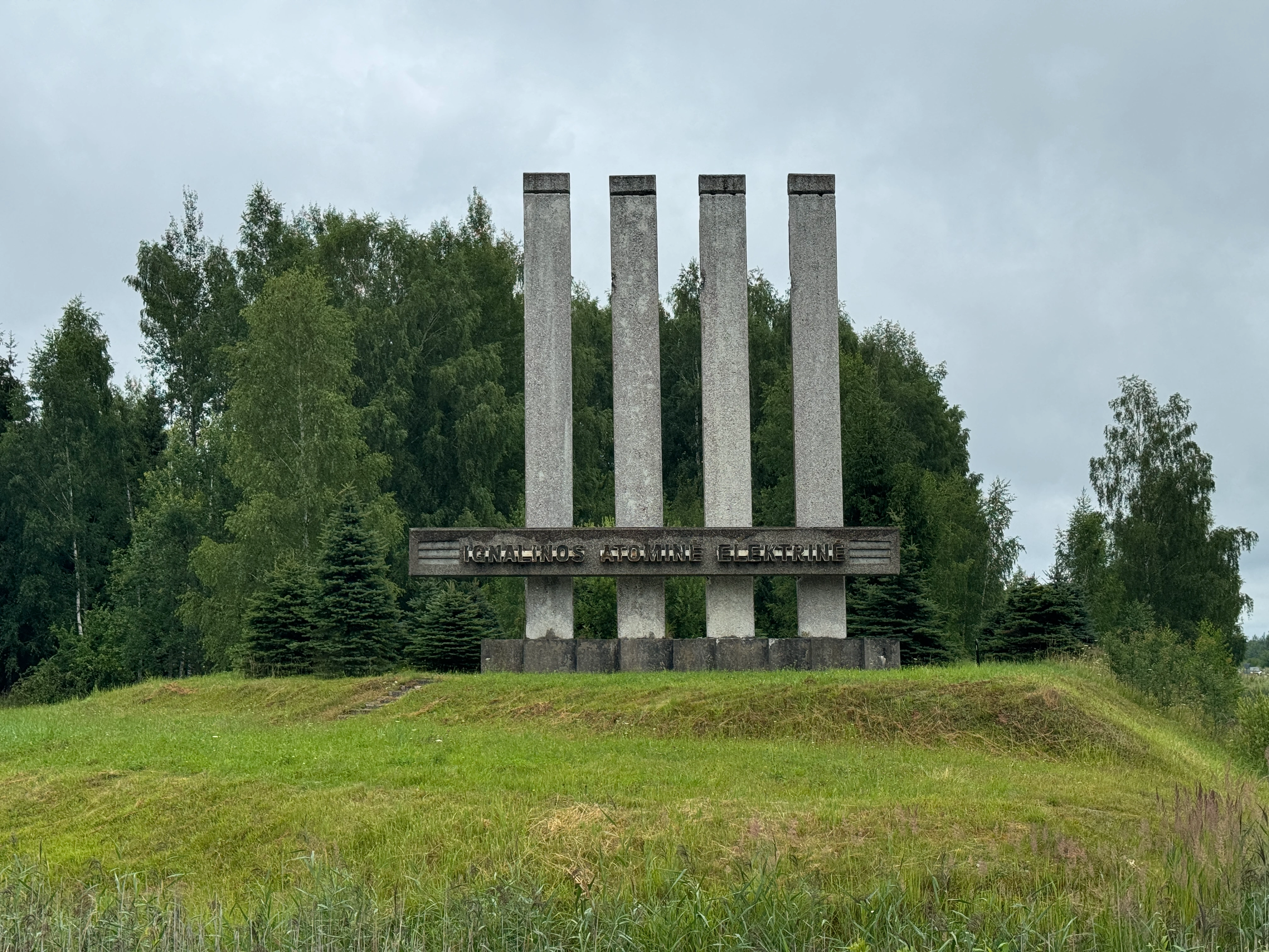 Close-up of the concrete brutalist entrance monument to the Ignalina Nuclear Power Plant in Visaginas, Lithuania, focusing on the horizontal crossbar with metal lettering spelling "IGNALINOS ATOMINĖ ELEKTRINĖ" across four vertical pillars.