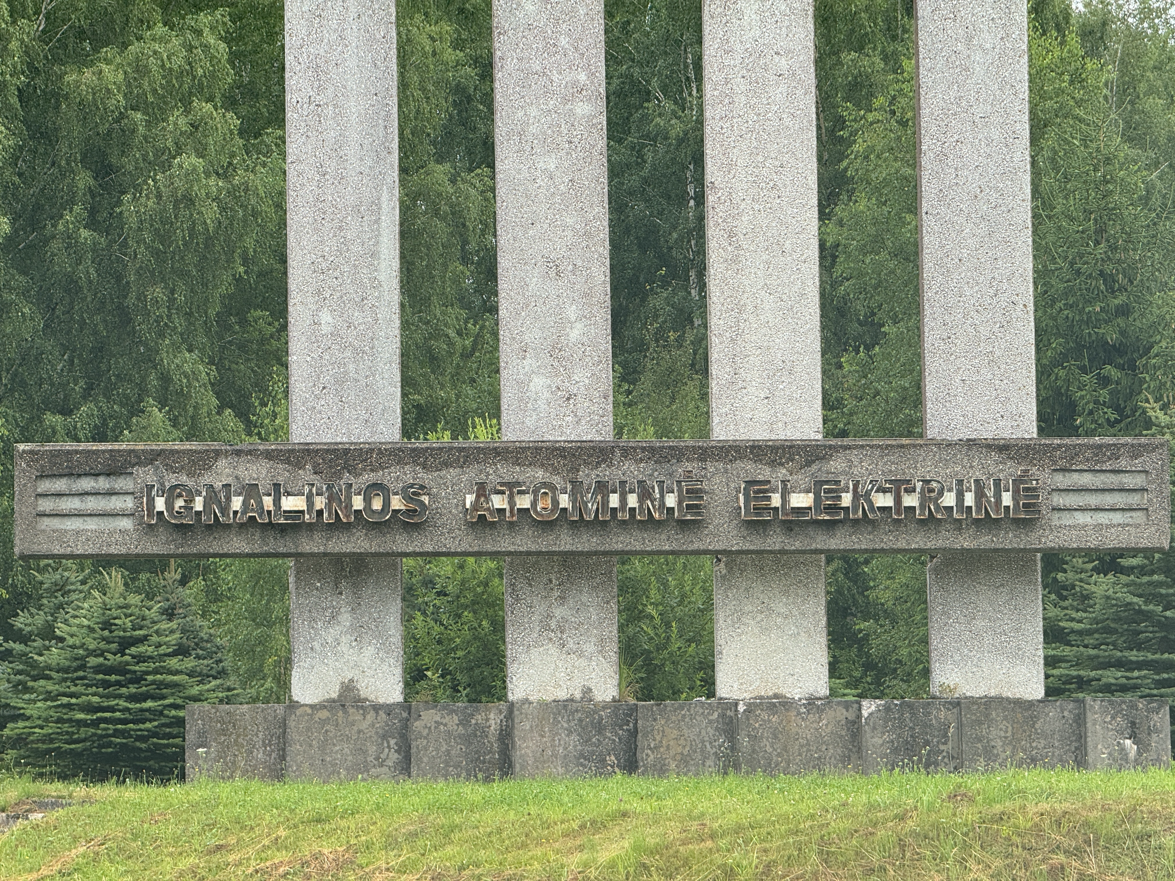 Close-up of the concrete brutalist entrance monument to the Ignalina Nuclear Power Plant in Visaginas, Lithuania, focusing on the horizontal crossbar with metal lettering spelling "IGNALINOS ATOMINĖ ELEKTRINĖ" across four vertical pillars.