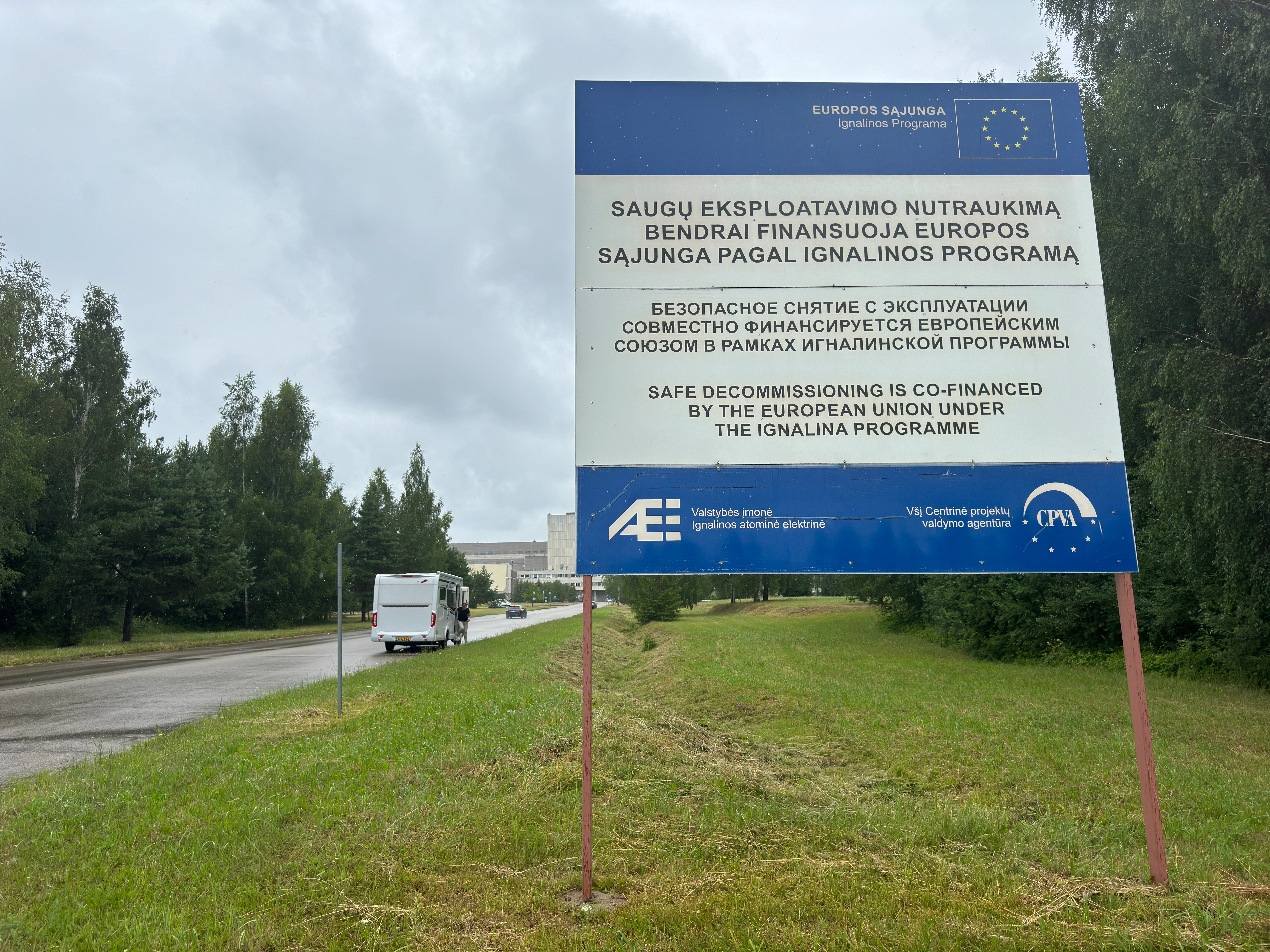 Large roadside sign near the Ignalina Nuclear Power Plant in Visaginas, Lithuania. The sign displays 'SAFE DECOMMISSIONING IS CO-FINANCED BY THE EUROPEAN UNION UNDER THE IGNALINA PROGRAMME' in Lithuanian, Russian, and English, with the power plant building visible in the distant background.