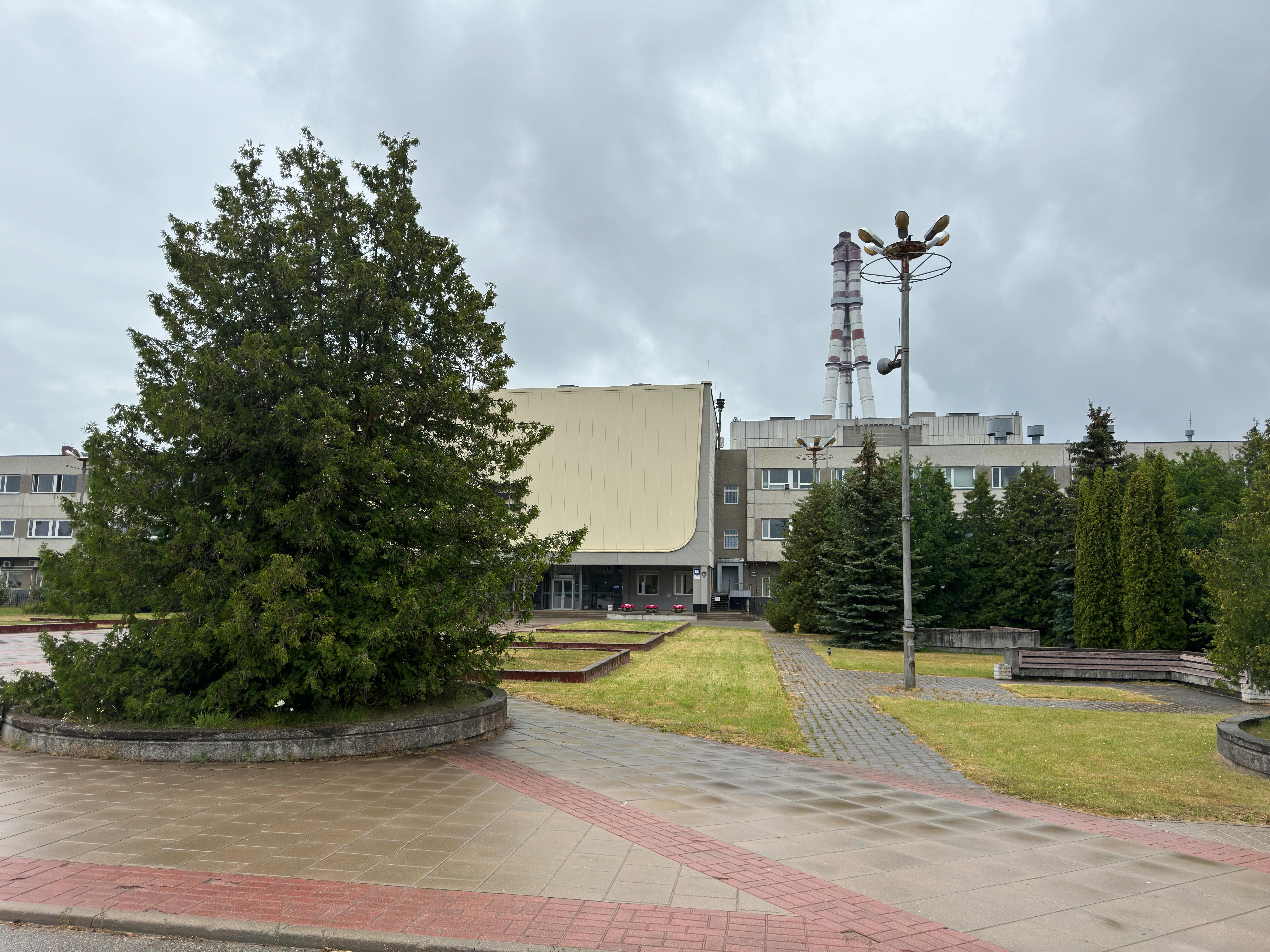 Exterior of the main administrative building at the Ignalina Nuclear Power Plant in Visaginas, Lithuania. The building features a large pale yellow overhanging facade with a dark 'AE' logo above the glass entrances. A paved plaza is in the foreground, and a striped ventilation stack is visible in the background under an overcast sky.