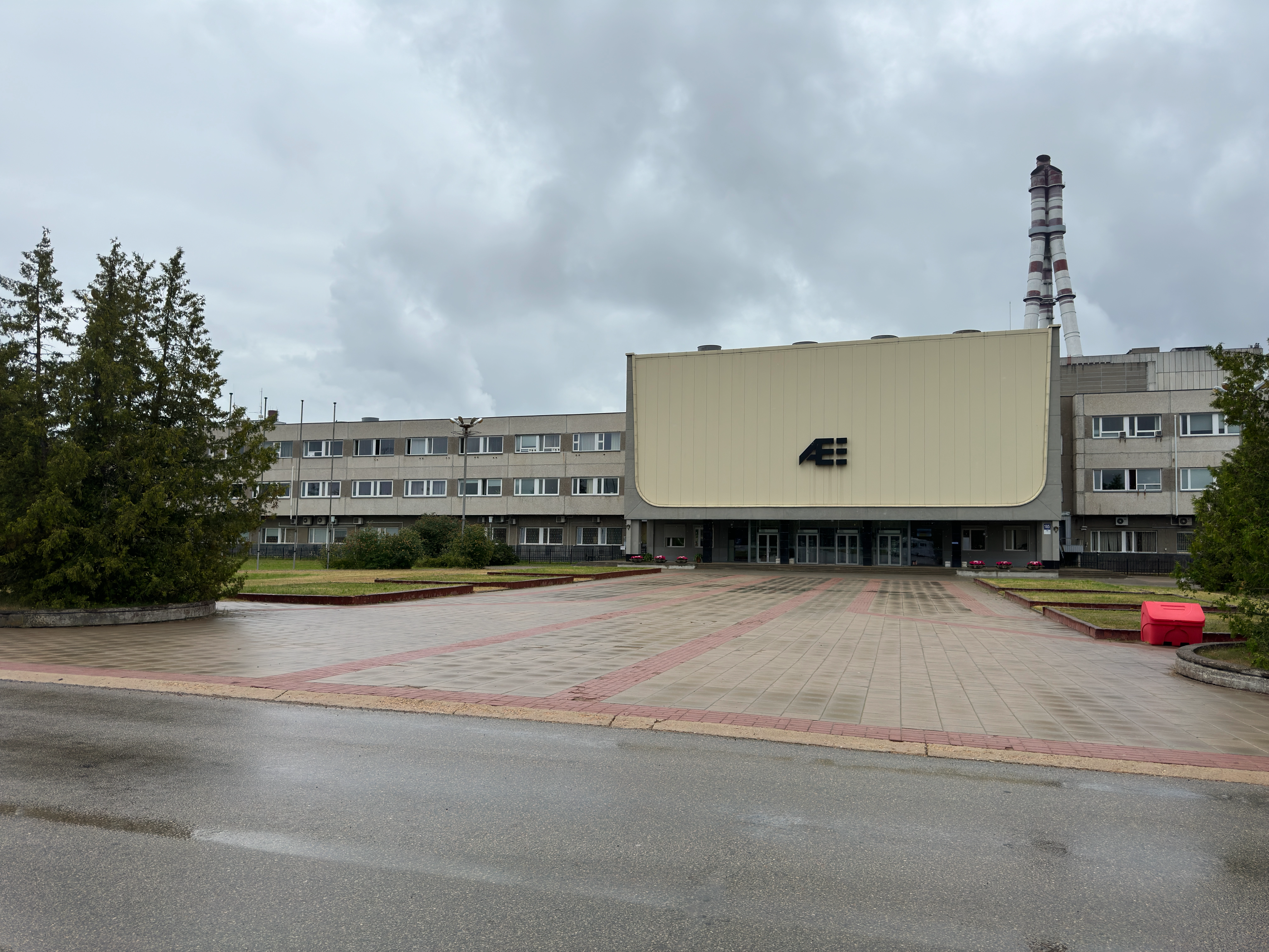 Exterior of the main administrative building at the Ignalina Nuclear Power Plant in Visaginas, Lithuania. The building features a large pale yellow overhanging facade with a dark 'AE' logo above the glass entrances. A paved plaza is in the foreground, and a striped ventilation stack is visible in the background under an overcast sky.