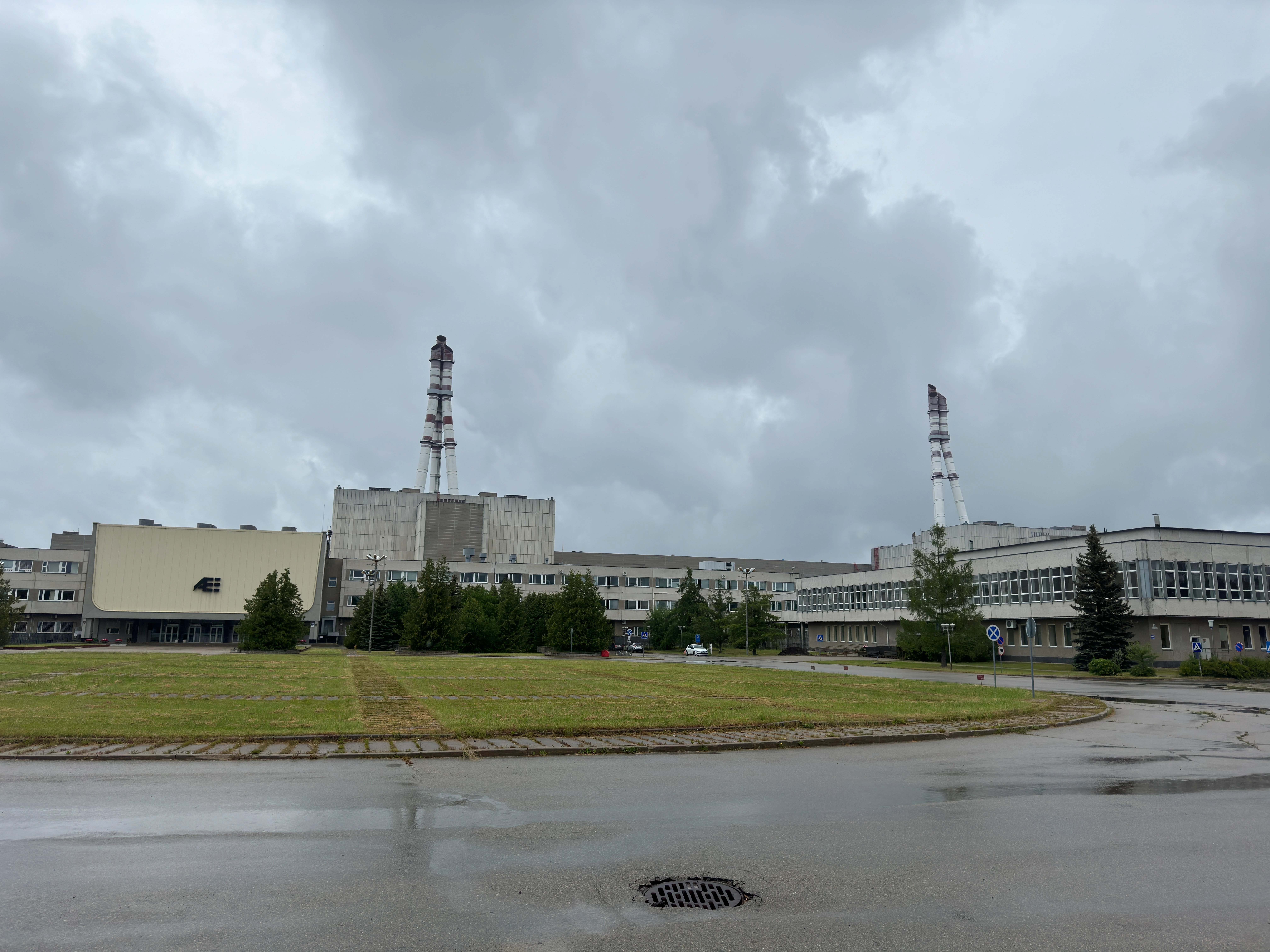 Wide exterior view of the Ignalina Nuclear Power Plant complex in Visaginas, Lithuania. The image shows the main administrative building with the 'AE' logo on the left, connected to large concrete reactor buildings featuring two tall, striped ventilation stacks against an overcast sky. A wet road and a grassy area are in the foreground.