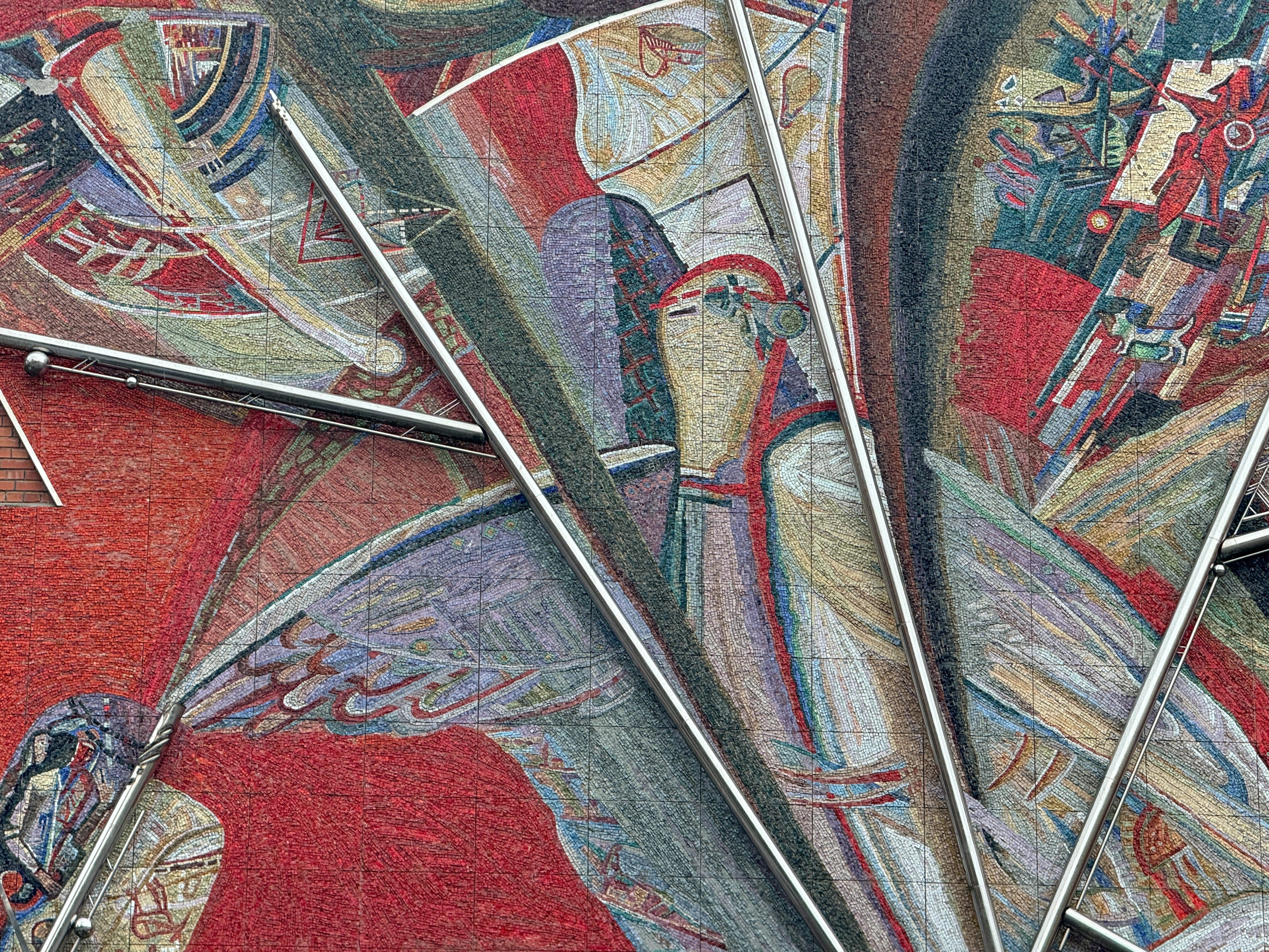 Close-up of a Soviet modernist mosaic on a building facade in Visaginas, Lithuania. The artwork features a textured red background with stylized, abstract figures in muted colors, intersected by protruding stainless steel rods and spheres.