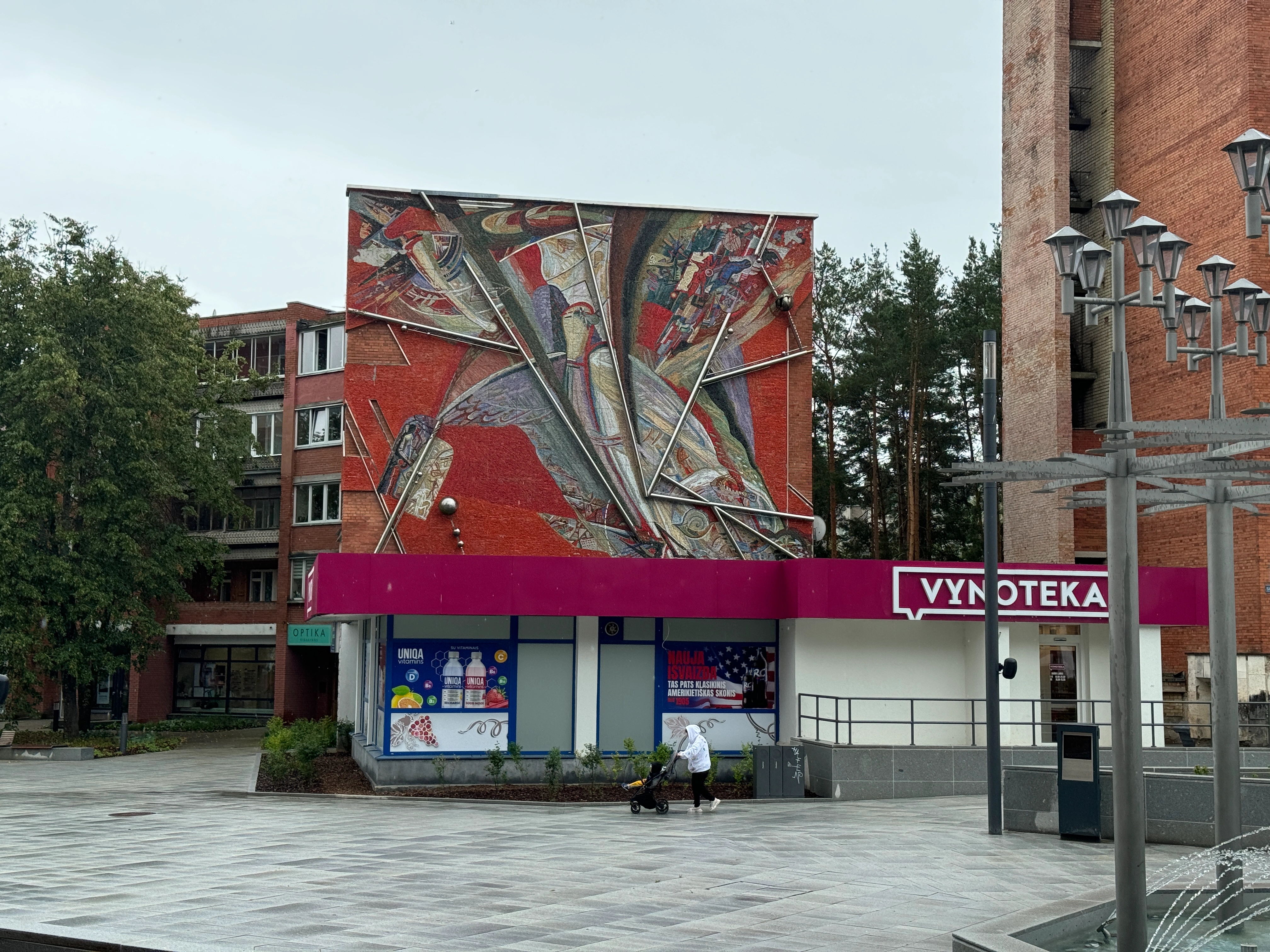 Wide view of a pedestrian square in Visaginas, Lithuania, featuring a red brick building with a large Soviet modernist mosaic on its upper facade. A Vynoteka store with a magenta awning is on the ground floor. A person pushes a stroller across the paved square.